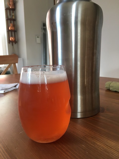 A plastic, stemless wine glass filled with a rosé-colored beer in front of a stainless steel 64 beer growler. They are on a wooden table. Background is white walls, a wooden chair back, and some general household decorations.
