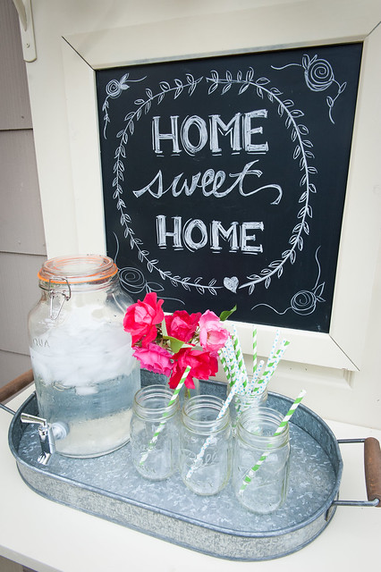 Upclose of the outdoor bar lower shelf with a Home Sweet Home chalkboard sign behind the beverage server and glasses