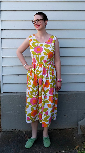 A woman stands in front of a weatherboard house. She wears a sleeveless fit and flare dress in a bright, Marimekko-style print and green flocked brogues.
