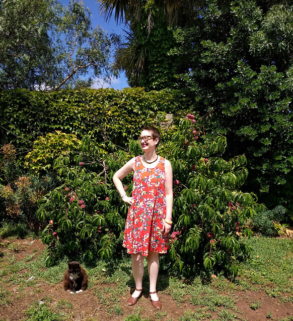 A woman stands in a garden. She wears a knee-length tent dress in an orange tropical floral print. A black and white cat sits next to her.
