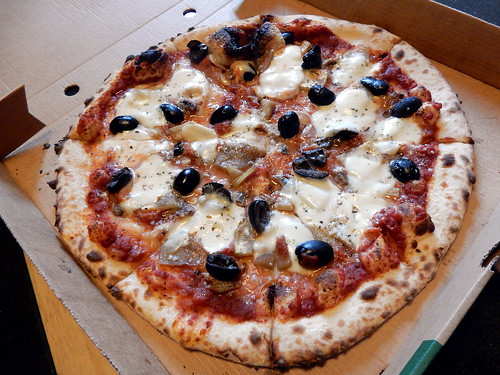 A pizza inside an open cardboard delivery box.  The pizza is topped with tomato sauce, black olives, and cheese.