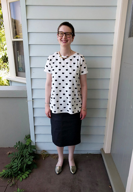 A woman stands in a veranda. She wears a white tee with black cat print, and a straight black denim skirt, with gold necklace and shoes.