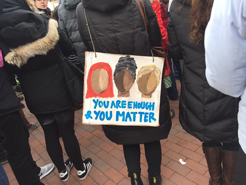 A protest sign, with three women’s faces painted on it. Beneath the faces, these words appear: “You are enough and you matter”