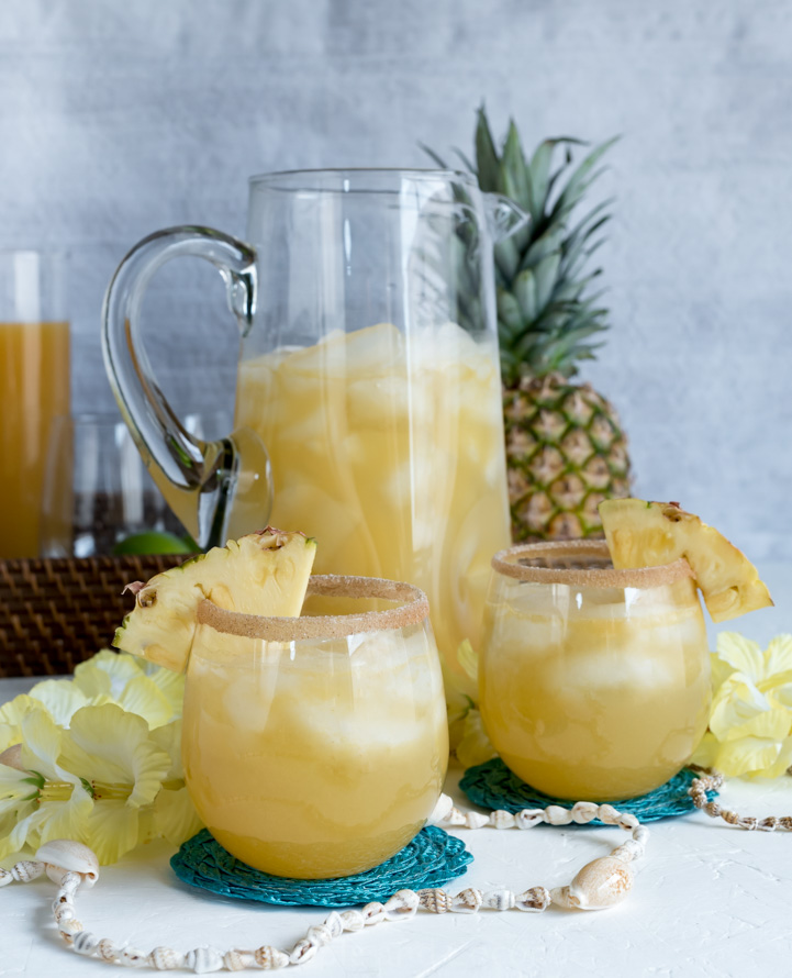 large pitcher of yellow rum punch cocktail, two smaller glasses of rum punch, whole pineapple, yellow flower lei, brown tray, shell necklace