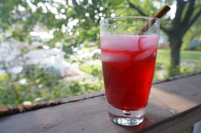 A glass of grown-up fruit punch on a porch rail, with a cinnamon stick leaning on the rim as a garnish