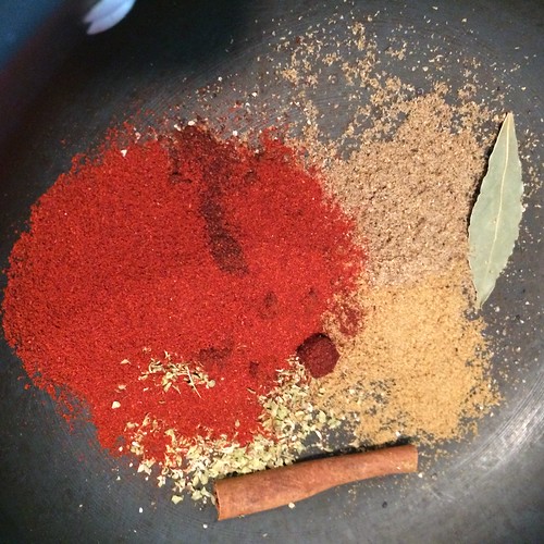 A colorful mix of spices and dried herbs in a pot. Smoked paprika, oregano, a bay leaf, a cinnamon stick, New Mexico red chile powder.