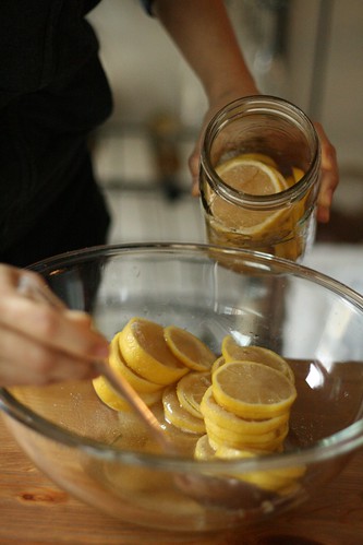 A close up of a woman's hands, spooning macerated lemons and their juce from a bowl into a wide-mouth quart canning jar.