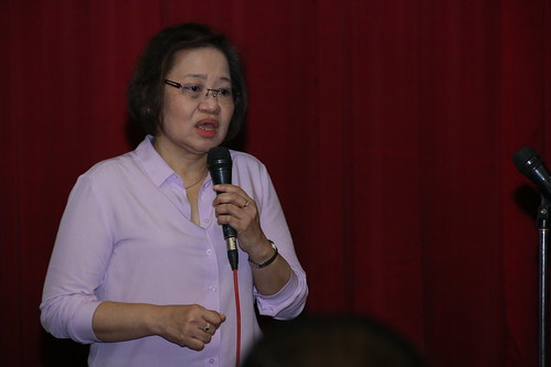 The image shows Dra. Alexis doing presentation during the seminar. She holds a microphone, wearing glasses and pink blouse. The background in red.