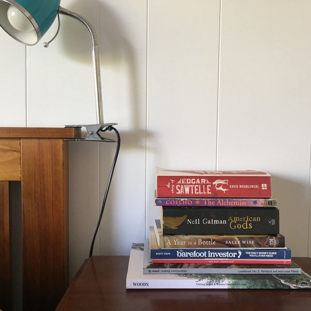 a stack of books and magazines on a bedside table with a reading lamp attached to the nearby bed