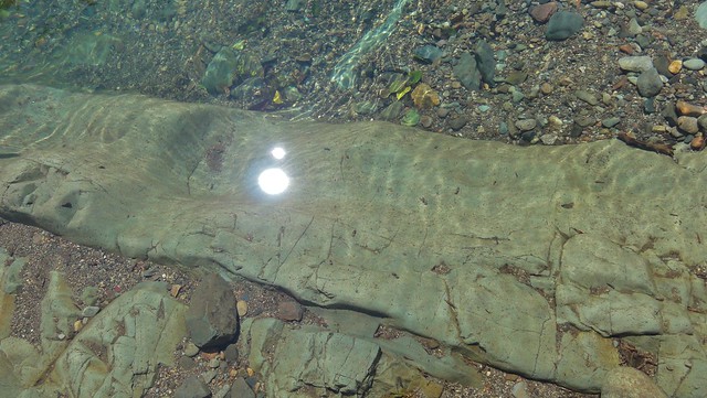 Image shows a basalt dyke under clear river water, framed by gravel. Sunlight is gleaming off the water above it.