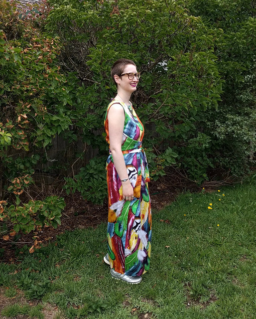 A woman stands in a garden. She wears a full length dress with ties at waist, in a colourful feather print, and silver runners.