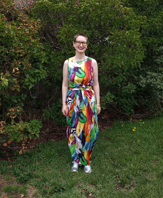 A woman stands in a garden. She wears a full length dress with ties at waist, in a colourful feather print, and silver runners.
