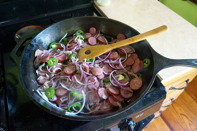 On a stovetop, a cast-iron skillet full of purple onion, bright green jalapeño, and disks of pink smoked sausage.