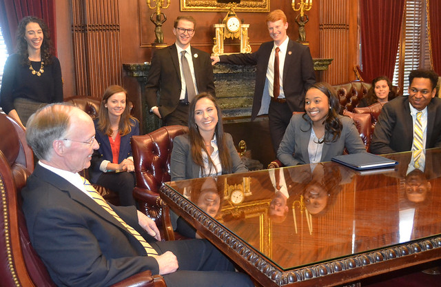 Alabama Gov. Robert Bentley sits at the head of a conference table while six students are sitting and standing around the table