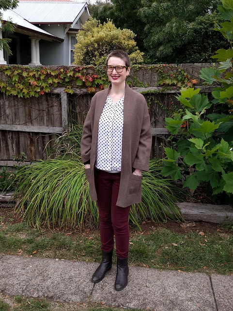 A woman stands against a garden fence. She wears a spotted button up shirt, maroon skinny jeans, brown boots and an oversized brown wool open coat.