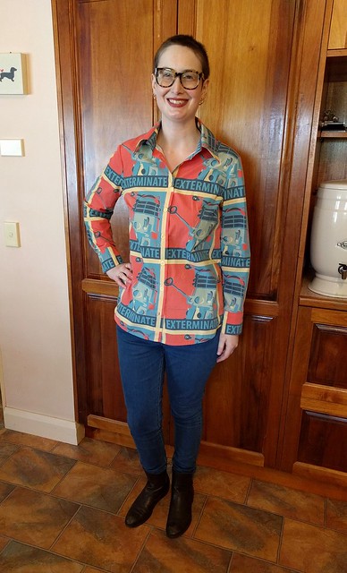 A woman wears a button up shirt with Dalek print, skinny blue jeans, and brown boots.