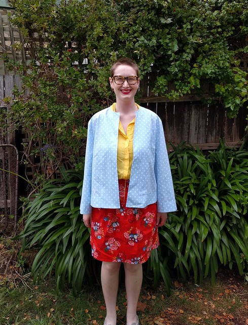 A woman stands against a garden fence. She wears a blue and white polka dot sun jacket, yellow shirt, and red floral print skirt.