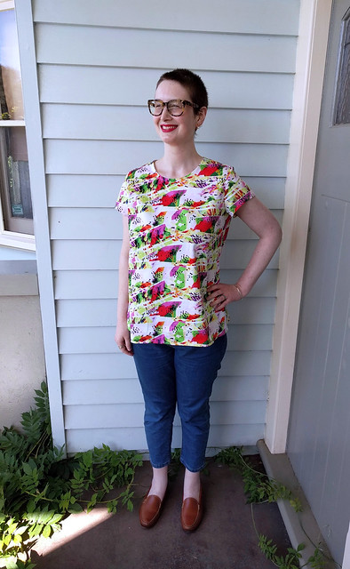 A woman stands in front of a weatherboard wall. She wears a colourful printed t-shirt, crop jeans, and leather loafers.
