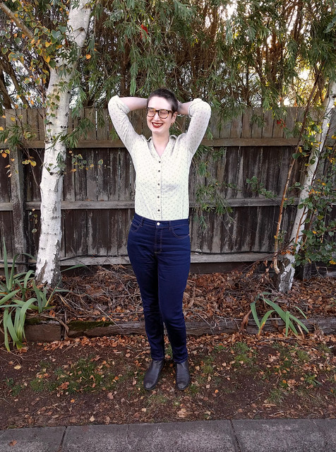 A woman stands in front of a garden fence. She wears dark blue, slim jeans and a button up shirt.
