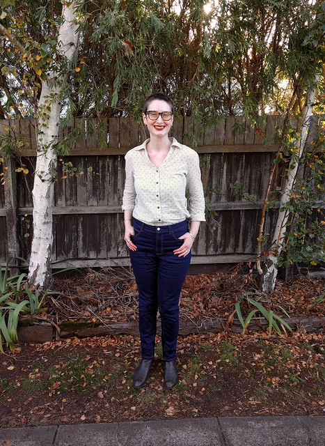 A woman stands in front of a garden fence. She wears dark blue, slim jeans and a button up shirt.