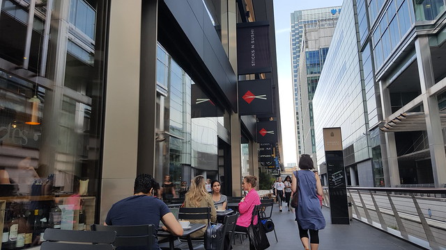 its a walkway flanked by tall buildings on both sides with people sitting out at tables. 'Sticks and Sushi' is one of the signs