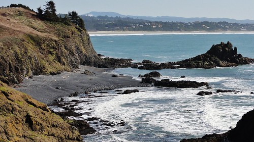 Image shows a brownish-gray rocky cliff curving around a dark gray cobble beach. A low arm of land fringed by tan sand stretches across the blue water in the distance.