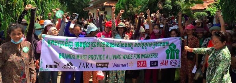 Garment workers hold up a sign reading "Brands must pay a living wage!" with the logos of various multinational brands lining the bottom of the banner 