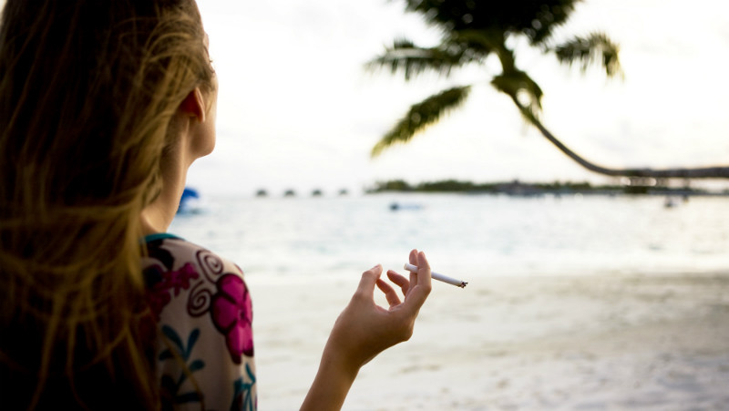 Image of woman in the foreground smoking on beach, palm tree and sea in background.