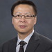 Portrait of Hou-Tong Chen wearing a black suit jacket and tie, with a black and white pinned strip shirt and wearing glasses