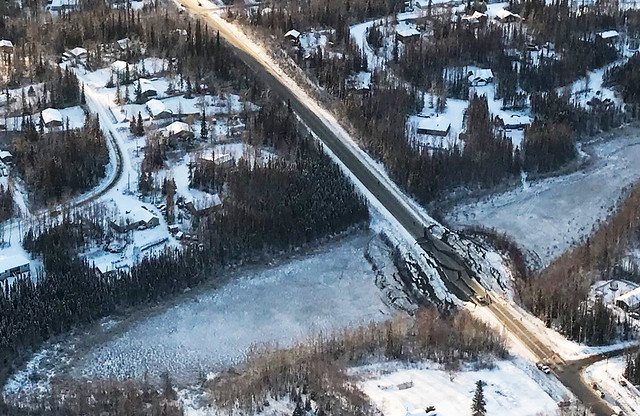 Image shows a snow-covered road from the air. The straight road goes through forested areas on either side of a depression that looks like a dry streambed. Within the bed, the road looks like an enormous wrecking ball was dropped on it, with huge cracks ripping it apart in a circular pattern. 