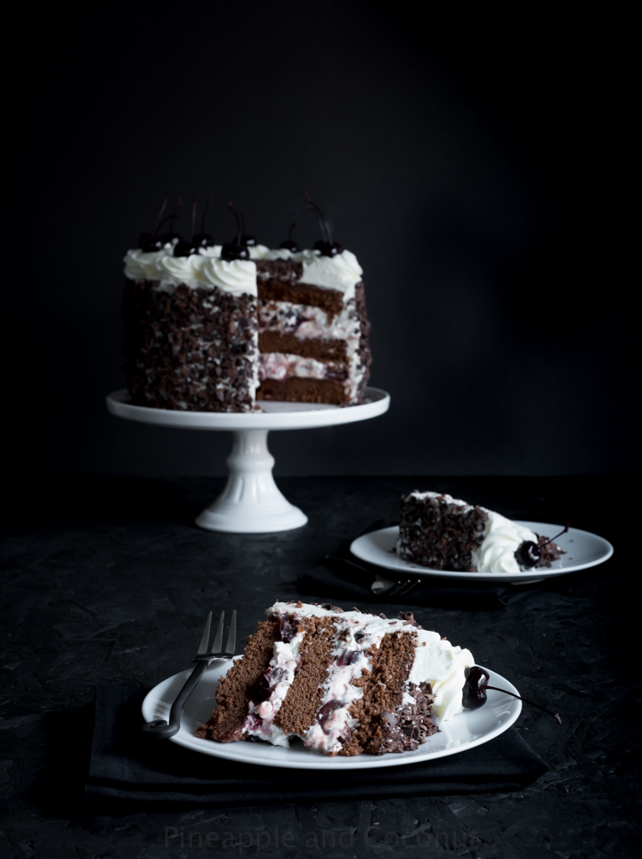 tall chocolate layer cake covered in chocolate curls on a white cake stand, filled and decorated with whipped cream and dark cherries two slices of cake on plates