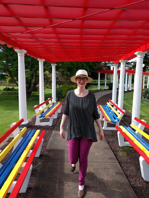 A woman stands under a pergola, wearing a draped knit tee, purple pants, and a panama hat.