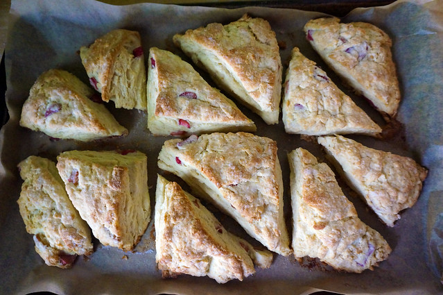 The same scone triangles, now baked. They sit slightly separated on a lined baking sheet, puffed and brown, the bits of rhubarb shining redly from between flaky layers.