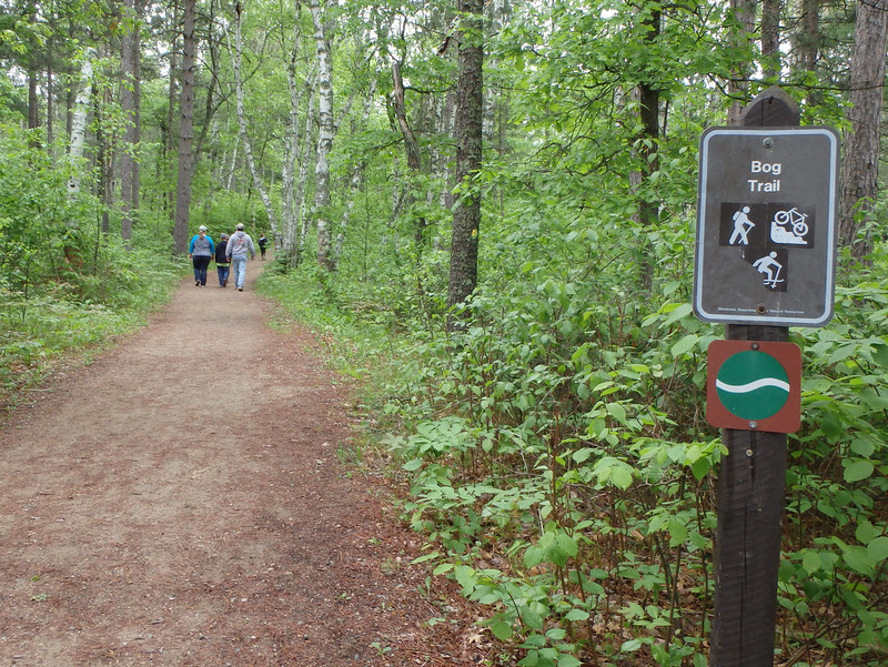 sign for the Bog Trail at the right, the unpaved path in the middle, with people walking away in the distance