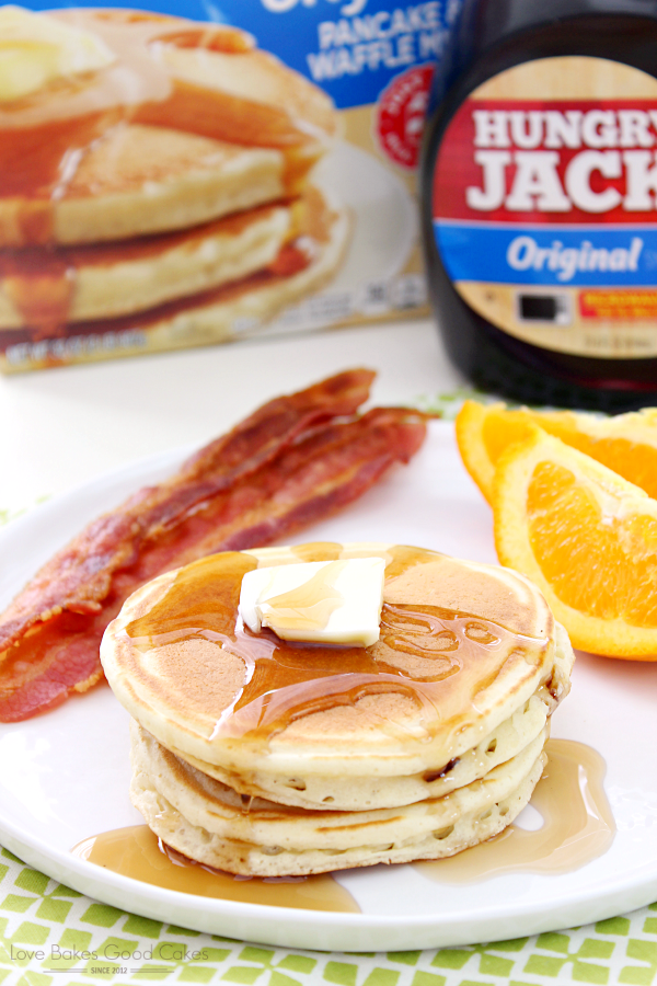 A stack of pancakes on a white plate with bacon slices and orange wedges and a bottle of hungry jack syrup.