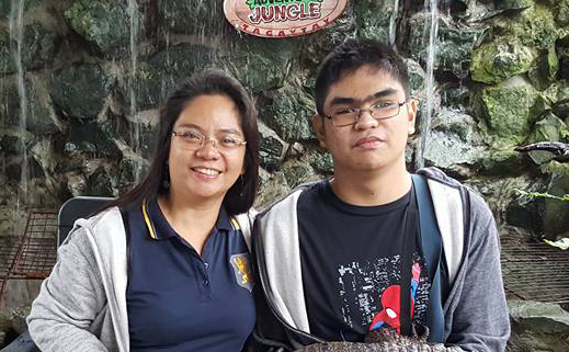 The image shows Ms. Joyla wearing ultrabmarine collared shirt and her son Juris wears black shirt with Spiderman design. Both of them wear eyeglasses and silver jacket.