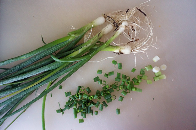 On a white cutting board, a pair of whole scallions, their tangled roots still slightly dirty, next to a pile of sliced scallion rounds, their colors forming a gradation from white to dark green that mirrors the whole scallions