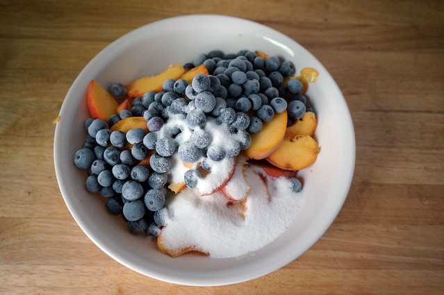 A tempting still life: in a wide ivory-colored bowl, a layer of bright yellow peaches, mostly covered by blueberries, frosted with freezer cold, the whole mount dusted with a light coat of sugar.