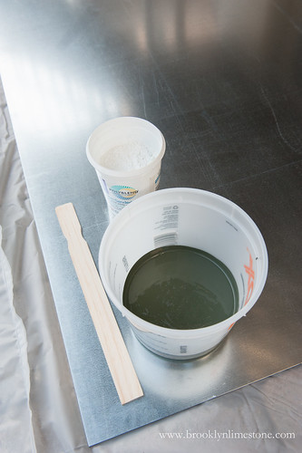 Plastic container holding outdoor paint, container of unsanded white grout with the top off and a stir stick
