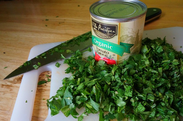 A small can of tomato sauce sits in the middle of a mound of chopped herbs, with a knife in the background, scattered bits of herbs on the blade. The picture makes no practical sense, but looks nice.