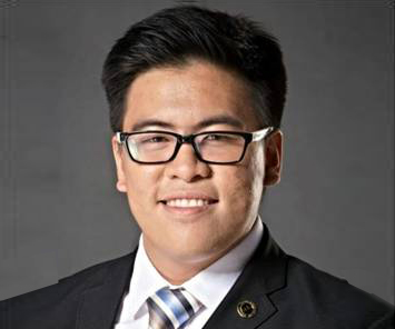 The image shows Mr. Dominic Cheoc wearing white polo with black and blue striped tie and black suit. He wears eyeglasses with his smile. His hair is almost straight up.