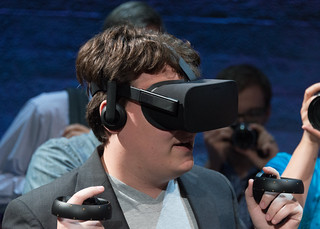 Palmer Luckey (Founder, Oculus) wearing Oculus Rift consumer version and holding up Oculus Touch prototype (Half Moon) controllers (closeup headshot / bust)